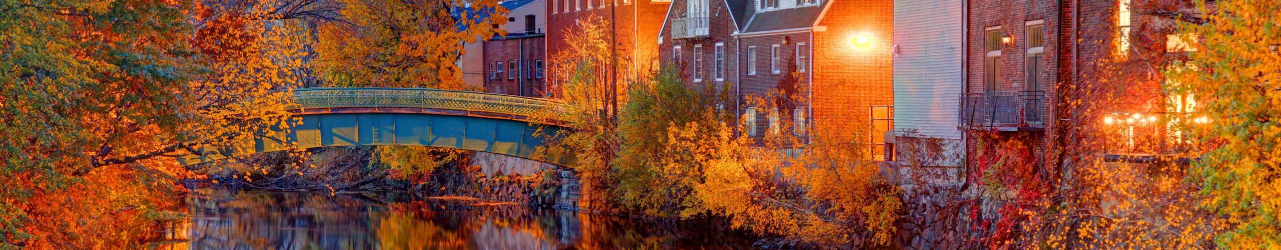 Colorful fall foliage in Massachusetts with a bridge over a river and building reflections in the water.