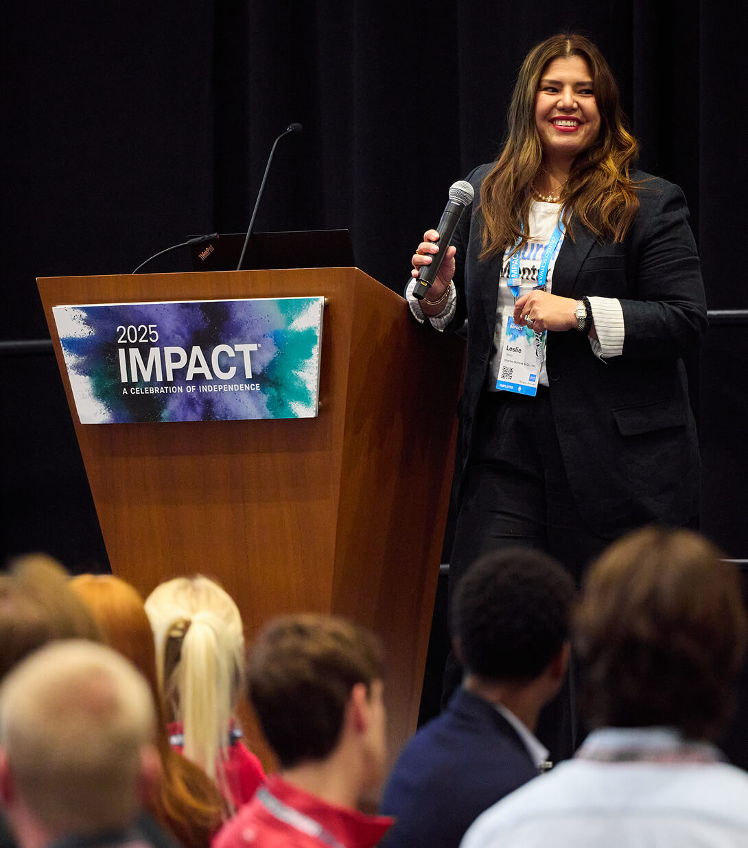 Smiling woman with a microphone next to a podium onstage at IMPACT 2025.