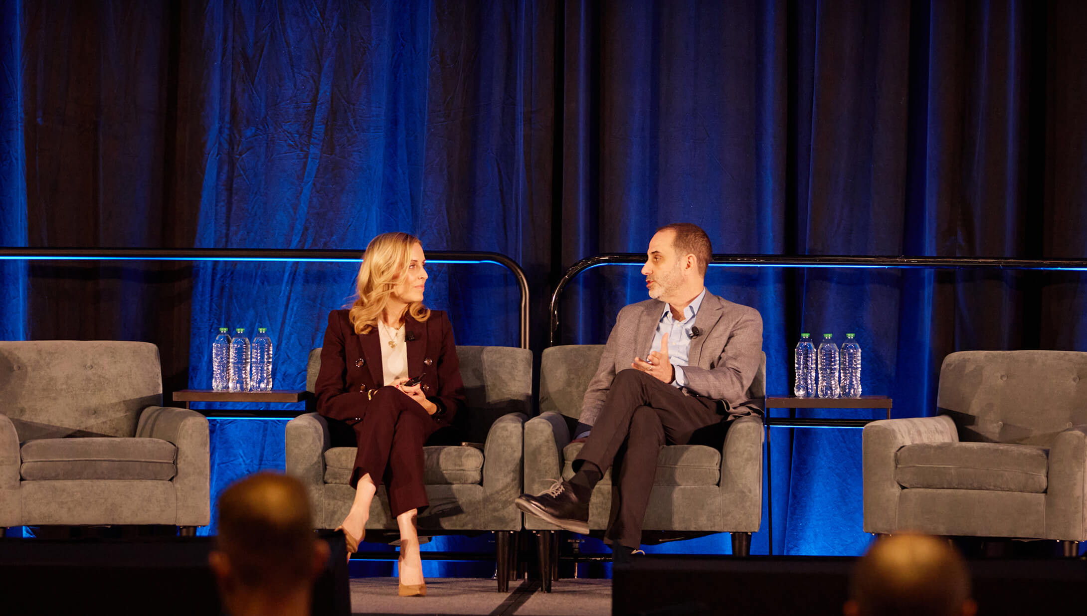 Seated man and woman conversing onstage in front of a blue drape at IMPACT 2025.