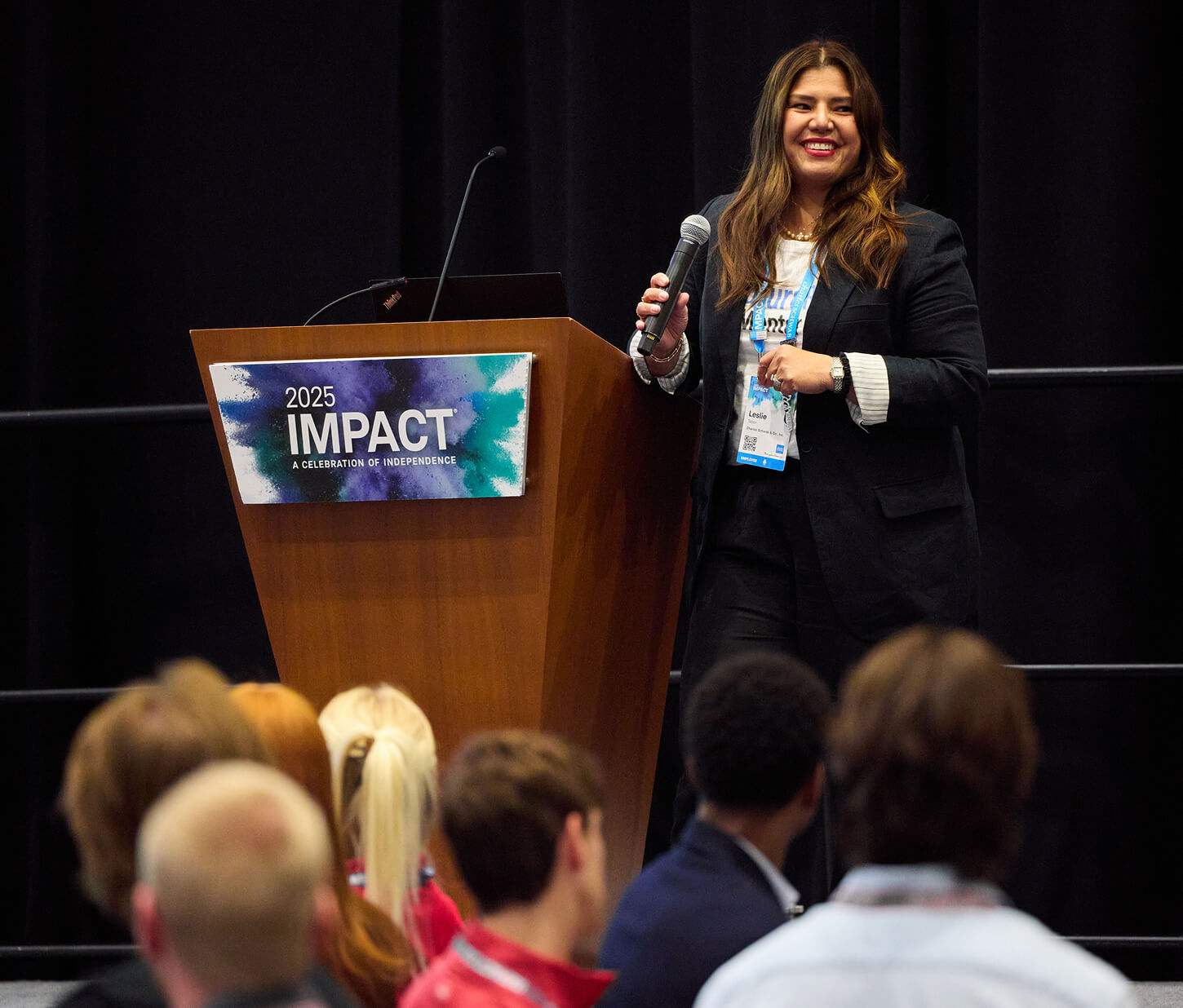Smiling woman with a microphone next to a podium onstage at IMPACT 2025.