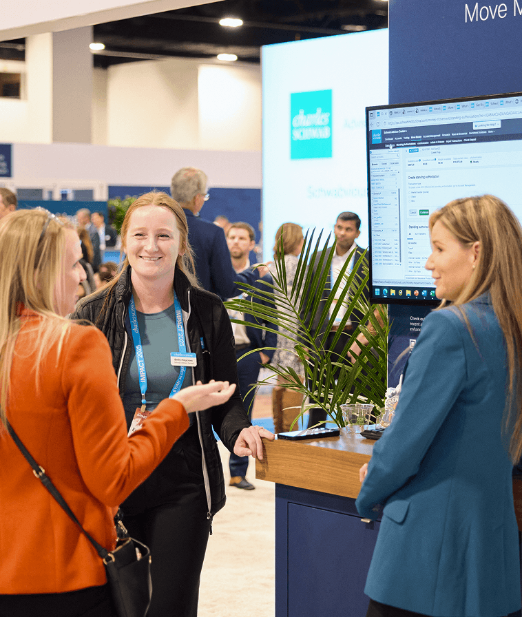 Three women conversing near a computer screen in The Exchange at IMPACT 2025.